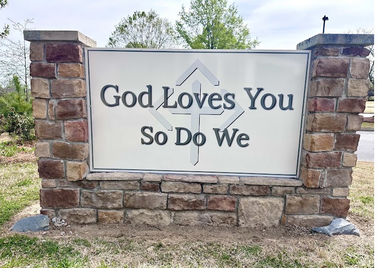 Harvest Church sign reading God Loves You So Do We, set in stone pillars at the church entrance